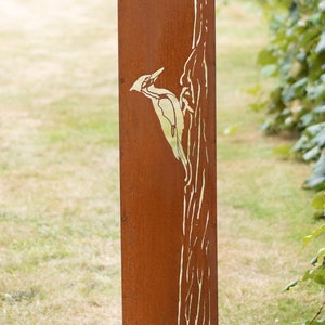 May include: A rectangular, rusty-brown metal garden stake featuring a cut-out design of a woodpecker perched on a tree trunk. The stake is supported by two thin metal legs, and is set in a grassy yard with green foliage in the background.