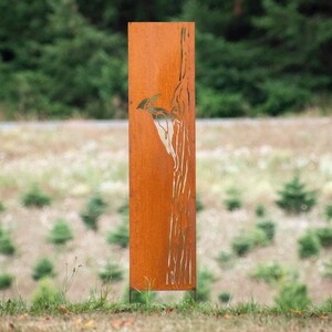 May include: A tall, rectangular, weathered metal sculpture with a bird design. The sculpture is a rusty orange color and stands in a grassy area with trees in the background. The artwork is a decorative outdoor element.