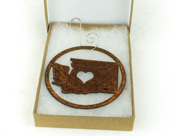 May include: A wooden ornament shaped like the state of Washington with a heart cutout in the center. The ornament is in a brown gift box with white tissue paper.