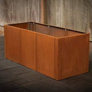May include: A rectangular, rust-colored planter box. The box is made of metal with vertical paneling and a weathered finish. The planter is empty and sits on a dark, textured surface, with a wooden fence in the background.
