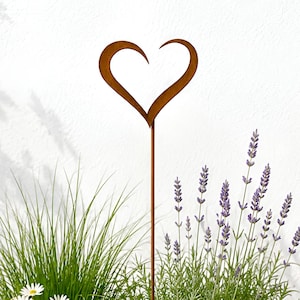 May include: A rust-colored metal heart-shaped garden stake stands upright in a large, round, light gray planter filled with green grass, purple lavender, small white daisy-like flowers, and a single smooth gray stone nestled in the soil.