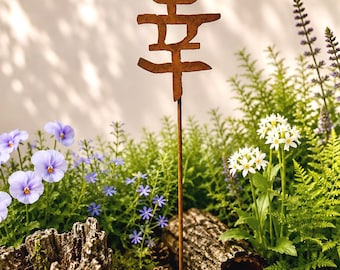 May include: A brown metal garden stake featuring the Japanese kanji character for happiness, positioned in a round ceramic planter filled with soil, moss, small pinecones, a piece of bark, green ferns, white flowers, and purple pansies.