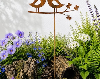 Op de afbeelding: Een roestkleurige metalen tuinpen met twee gestileerde vogels op een tak met kleine blaadjes. De pen staat in een ronde keramische plantenbak gevuld met paarse viooltjes, groene varens, mos, een dennenappel en stukken hout.