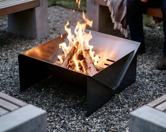 May include: A square, black metal fire pit with an angular, geometric design containing a bright, flickering wood fire. The fire pit features side panels with horizontal ventilation slots and sits on a gravel surface near wooden benches.