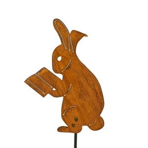 May include: A rusty brown metal garden stake featuring a rabbit reading a book. The rabbit is depicted in profile, with its ears perked up and its eyes focused on the book. The stake is designed for outdoor use.