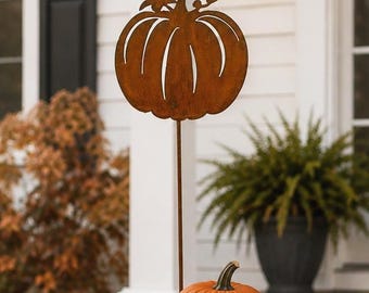 May include: A rust-colored metal pumpkin garden stake stands behind a hay bale. A real orange pumpkin, a white pumpkin, a striped gourd, and a small orange pumpkin sit near a wooden sign with the text hello FALL and a leaf graphic.