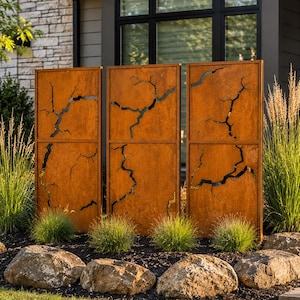 May include: Three rectangular, rust-colored metal privacy screens featuring a jagged, lightning-bolt-style cutout pattern. The panels are arranged vertically in a row, standing upright in a garden bed with dark mulch, large rocks, and tall grasses.