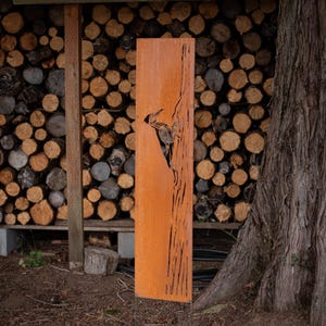 May include: A tall, rectangular, rusty-orange metal art piece featuring a cut-out design of a bird, likely a woodpecker. The artwork stands upright against a backdrop of stacked firewood and a large tree trunk.