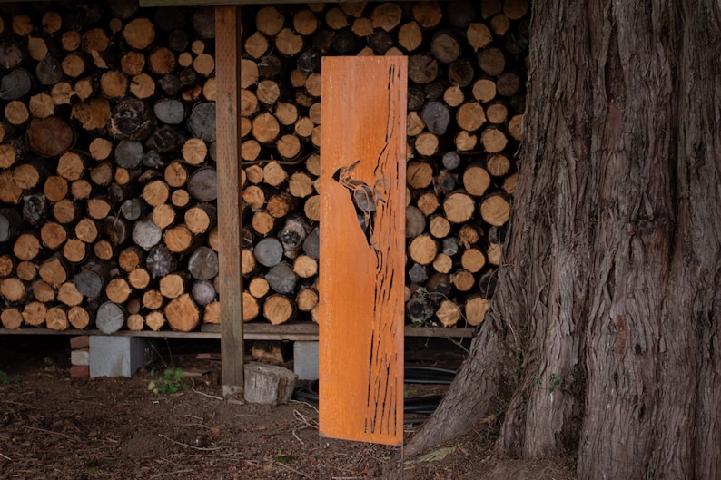 May include: A tall, rectangular, rusty-orange metal art piece featuring a cut-out design of a bird, likely a woodpecker. The artwork stands upright against a backdrop of stacked firewood and a large tree trunk.