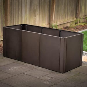 May include: A rectangular, dark brown metal planter box. The planter is made of several vertical panels and sits on a dark gray stone patio. A wooden fence and green grass are visible in the background.