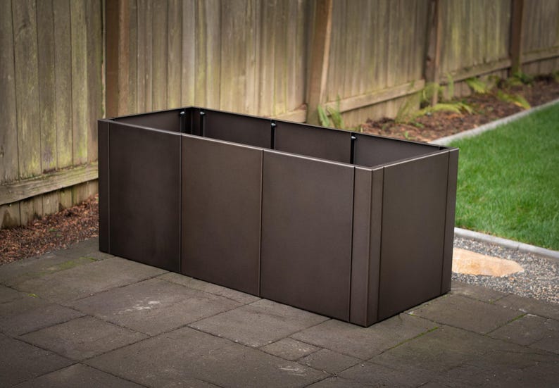 May include: A rectangular, dark brown metal planter box. The planter is made of several vertical panels and sits on a dark gray stone patio. A wooden fence and green grass are visible in the background.