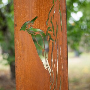 May include: A weathered, rectangular metal panel with a cut-out design of a woodpecker perched on a tree branch. The panel has a rusty brown color, and the background is blurred green foliage.