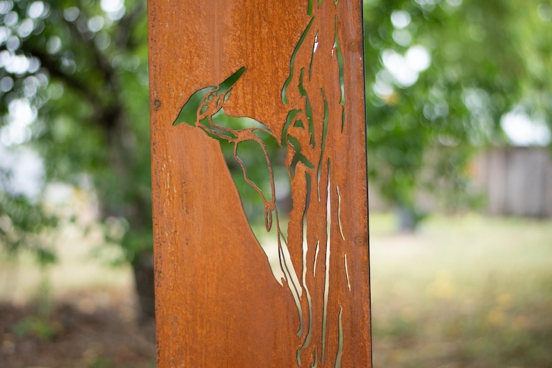 May include: A weathered, rectangular metal panel with a cut-out design of a woodpecker perched on a tree branch. The panel has a rusty brown color, and the background is blurred green foliage.