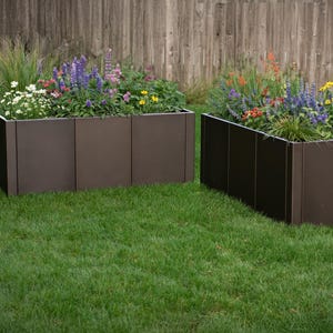 May include: Two dark brown rectangular raised garden beds filled with colorful flowers and greenery. The beds are on a green lawn, with a wooden fence in the background. The flowers include purple, yellow, and orange blooms.