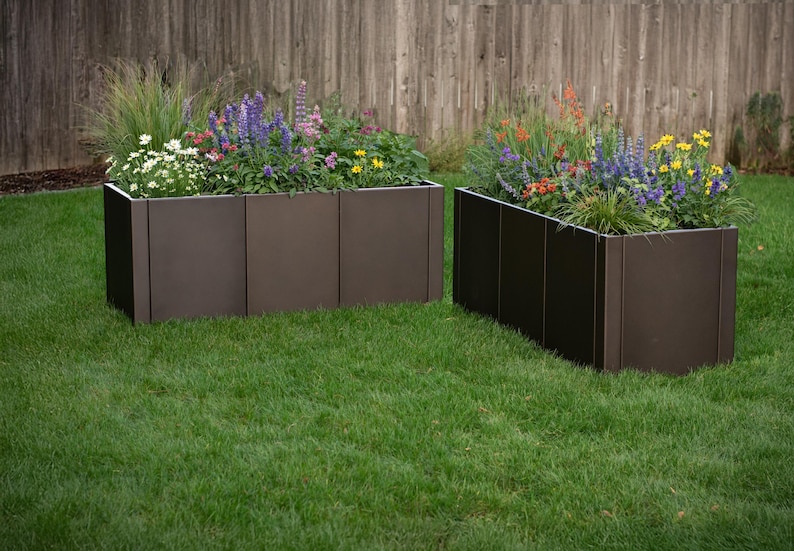 May include: Two dark brown rectangular raised garden beds filled with colorful flowers and greenery. The beds are on a green lawn, with a wooden fence in the background. The flowers include purple, yellow, and orange blooms.