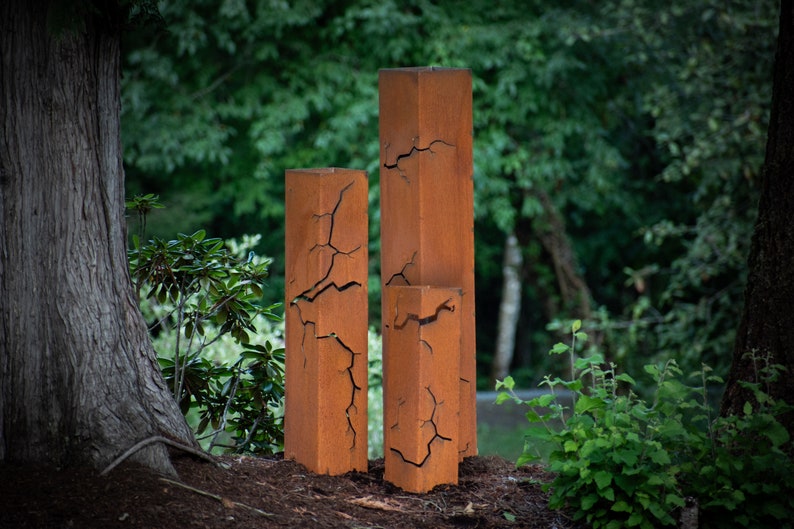 May include: Three tall, rectangular, rusty metal sculptures with cracks throughout the surface. The sculptures are standing on a bed of brown mulch.