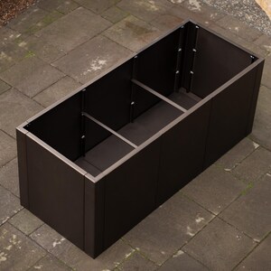 May include: A rectangular, dark brown planter box with a silver trim. The planter is divided into three sections and sits on a stone patio. The box is empty and ready for plants.
