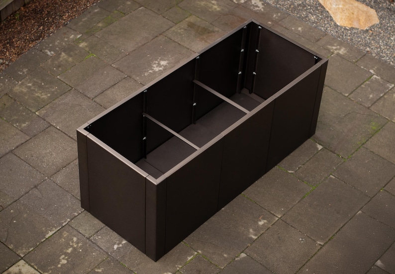 May include: A rectangular, dark brown planter box with a silver trim. The planter is divided into three sections and sits on a stone patio. The box is empty and ready for plants.