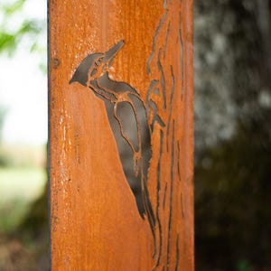 May include: A weathered, rectangular metal panel with a cut-out design of a woodpecker perched on a tree branch. The panel has a rusty orange color, with the bird and tree silhouette in dark brown.