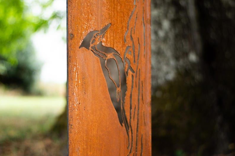 May include: A weathered, rectangular metal panel with a cut-out design of a woodpecker perched on a tree branch. The panel has a rusty orange color, with the bird and tree silhouette in dark brown.