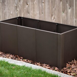 May include: A rectangular, dark brown metal planter box. The planter is empty and sits on a bed of fallen leaves and grass, with a wooden fence in the background. The planter is approximately 48 inches long.