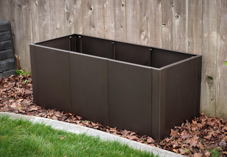 May include: A rectangular, dark brown metal planter box. The planter is empty and sits on a bed of fallen leaves and grass, with a wooden fence in the background. The planter is approximately 48 inches long.