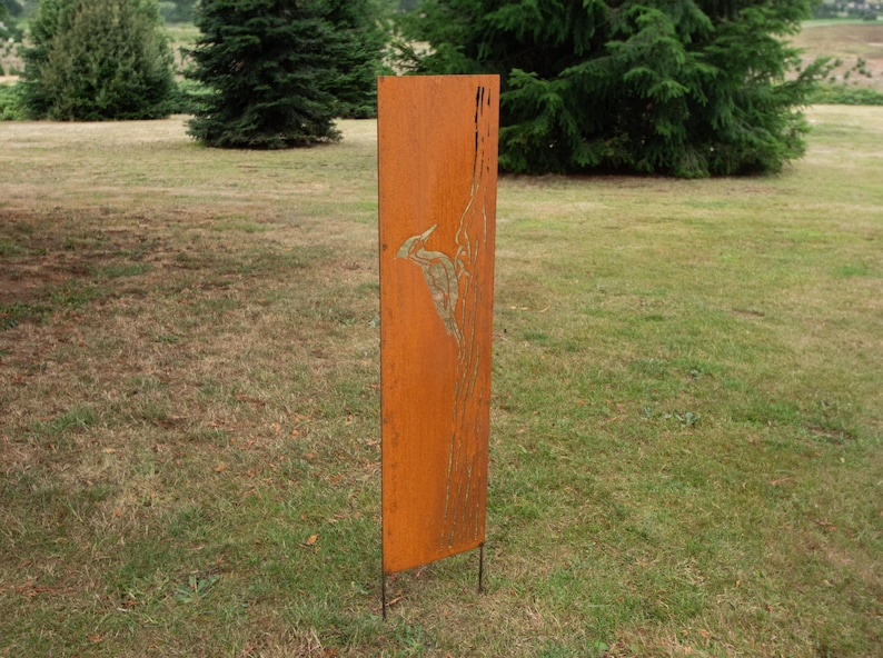 May include: A tall, rectangular, weathered metal garden art piece with a bird design. The artwork is a rusty orange color and stands on two metal legs, placed in a grassy outdoor setting.