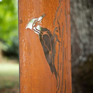 May include: A weathered, rectangular metal panel with a cut-out design of a woodpecker perched on a tree. The panel has a rusty orange color and is set against a blurred green background.