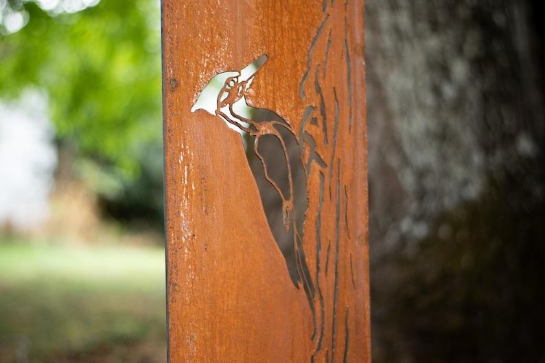 May include: A weathered, rectangular metal panel with a cut-out design of a woodpecker perched on a tree. The panel has a rusty orange color and is set against a blurred green background.