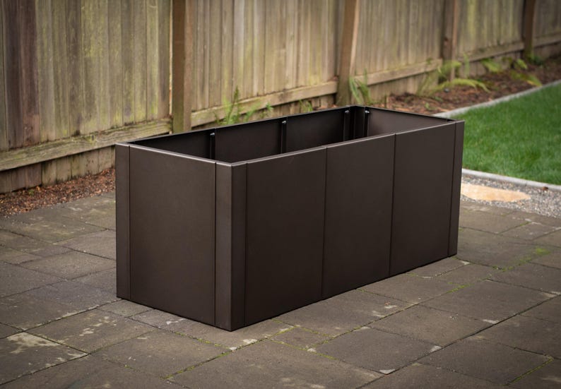 May include: A rectangular, dark brown planter box made of metal. The planter is empty and sits on a brick patio, with a wooden fence and green grass in the background. The box is approximately 36 inches long.