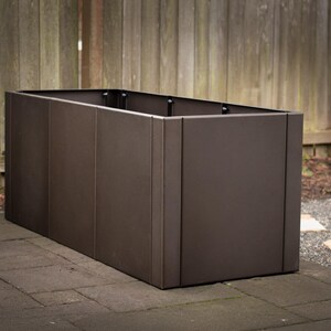 May include: A rectangular, dark brown planter box made of metal. The box is placed on a stone patio with a wooden fence in the background. The planter has a modern, minimalist design.