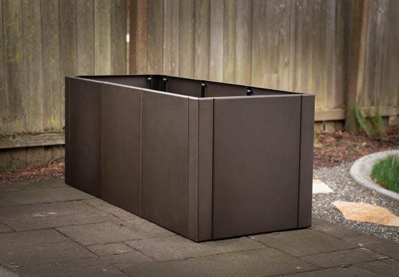 May include: A rectangular, dark brown planter box made of metal. The box is placed on a stone patio with a wooden fence in the background. The planter has a modern, minimalist design.