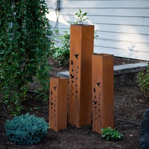 May include: Three tall, rust-colored metal planters with cut-out designs of birds and flowers. The planters are arranged in a row from tallest to shortest.