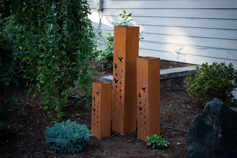 May include: Three tall, rust-colored metal planters with cut-out designs of birds and flowers. The planters are arranged in a row from tallest to shortest.