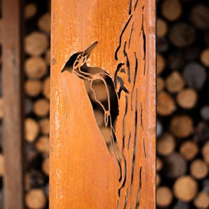May include: A weathered, orange-brown metal panel features a cut-out design of a woodpecker perched on a tree trunk. The background shows a blurred arrangement of cut logs, suggesting a rustic, outdoor theme.