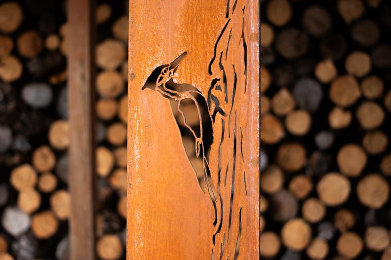 May include: A weathered, orange-brown metal panel features a cut-out design of a woodpecker perched on a tree trunk. The background shows a blurred arrangement of cut logs, suggesting a rustic, outdoor theme.