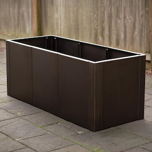 May include: A rectangular, dark brown planter box with a white trim around the top edge. The planter is made of vertical panels and sits on a stone patio. The background features a wooden fence.