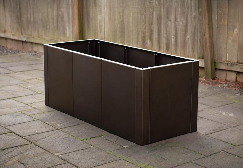 May include: A rectangular, dark brown planter box with a white trim around the top edge. The planter is made of vertical panels and sits on a stone patio. The background features a wooden fence.