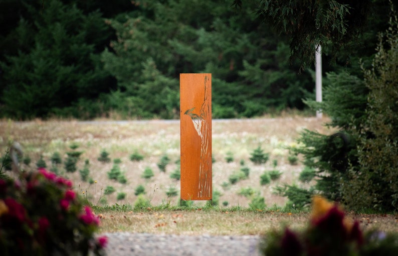 May include: A tall, rectangular, weathered metal sculpture with a bird design. The sculpture is a rusty orange color and stands in a grassy area with trees in the background. The artwork is a decorative outdoor element.