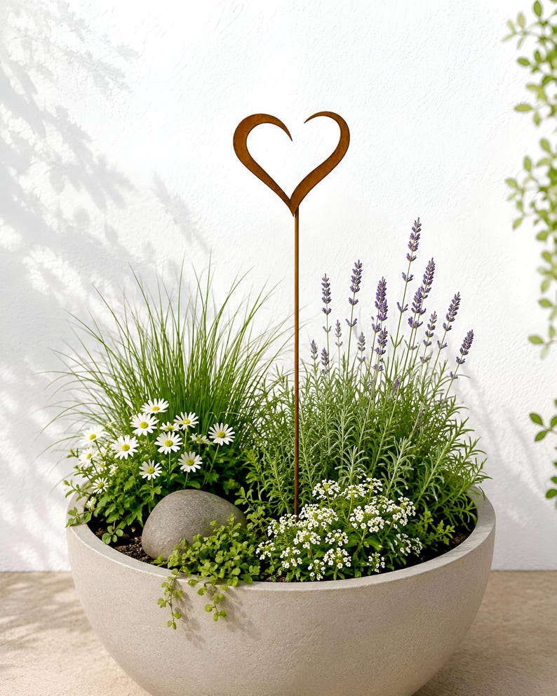 May include: A rust-colored metal heart-shaped garden stake stands upright in a large, round, light gray planter filled with green grass, purple lavender, small white daisy-like flowers, and a single smooth gray stone nestled in the soil.