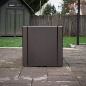 May include: A large, square, dark gray planter box. The planter has a modern design with vertical lines on the sides. It is sitting on a stone patio in front of a green lawn and wooden fence.