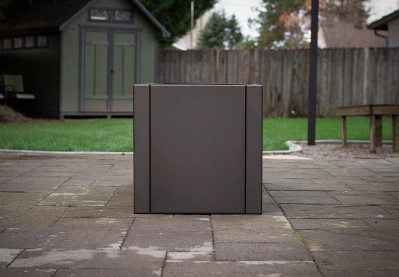 May include: A large, square, dark gray planter box. The planter has a modern design with vertical lines on the sides. It is sitting on a stone patio in front of a green lawn and wooden fence.