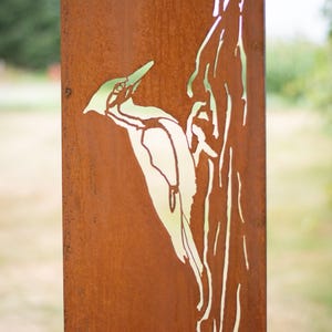 May include: A weathered metal sign featuring a cut-out design of a woodpecker perched on a tree branch. The sign has a rusty brown finish with a green and white woodpecker silhouette. The background is blurred, suggesting an outdoor setting.