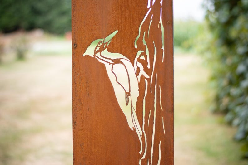 May include: A weathered metal sign featuring a cut-out design of a woodpecker perched on a tree branch. The sign has a rusty brown finish with a green and white woodpecker silhouette. The background is blurred, suggesting an outdoor setting.