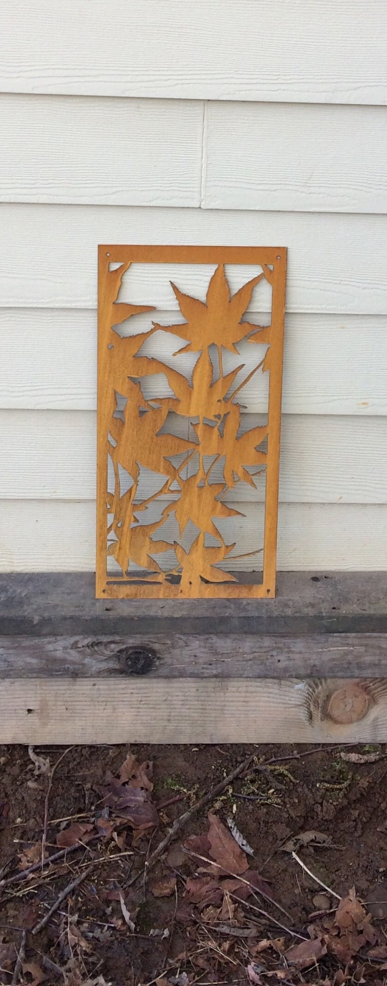 Op de afbeelding: Een roestige metalen wanddecoratie met een uitgesneden ontwerp van esdoornbladeren. De decoratie is gemonteerd op een witte houten muur.