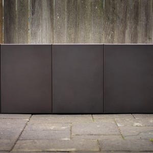 May include: A dark brown, rectangular outdoor planter box with three sections. The planter is made of a solid material and sits on a stone patio. A weathered wooden fence is in the background.