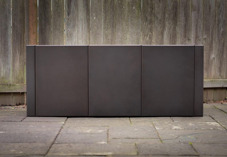 May include: A dark brown, rectangular outdoor planter box with three sections. The planter is made of a solid material and sits on a stone patio. A weathered wooden fence is in the background.