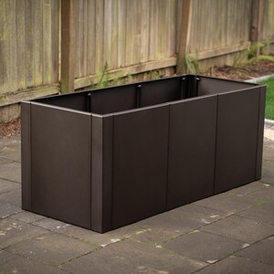 May include: A rectangular, dark brown planter box made of metal. The planter is empty and sits on a brick patio, with a wooden fence and grass in the background. The box is approximately 36 inches long.