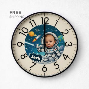 May include: A round wall clock with a black frame and a cream-colored face. The clock features a cartoon illustration of a baby astronaut on a planet, holding a ray gun. The clock hands are black.