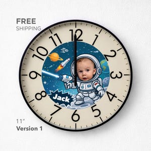 May include: A round wall clock with a black frame and a cream-colored face featuring black numerals. The center displays a space-themed illustration of an astronaut with a child's face, a rocket, planets, and the name Jack in a speech bubble.